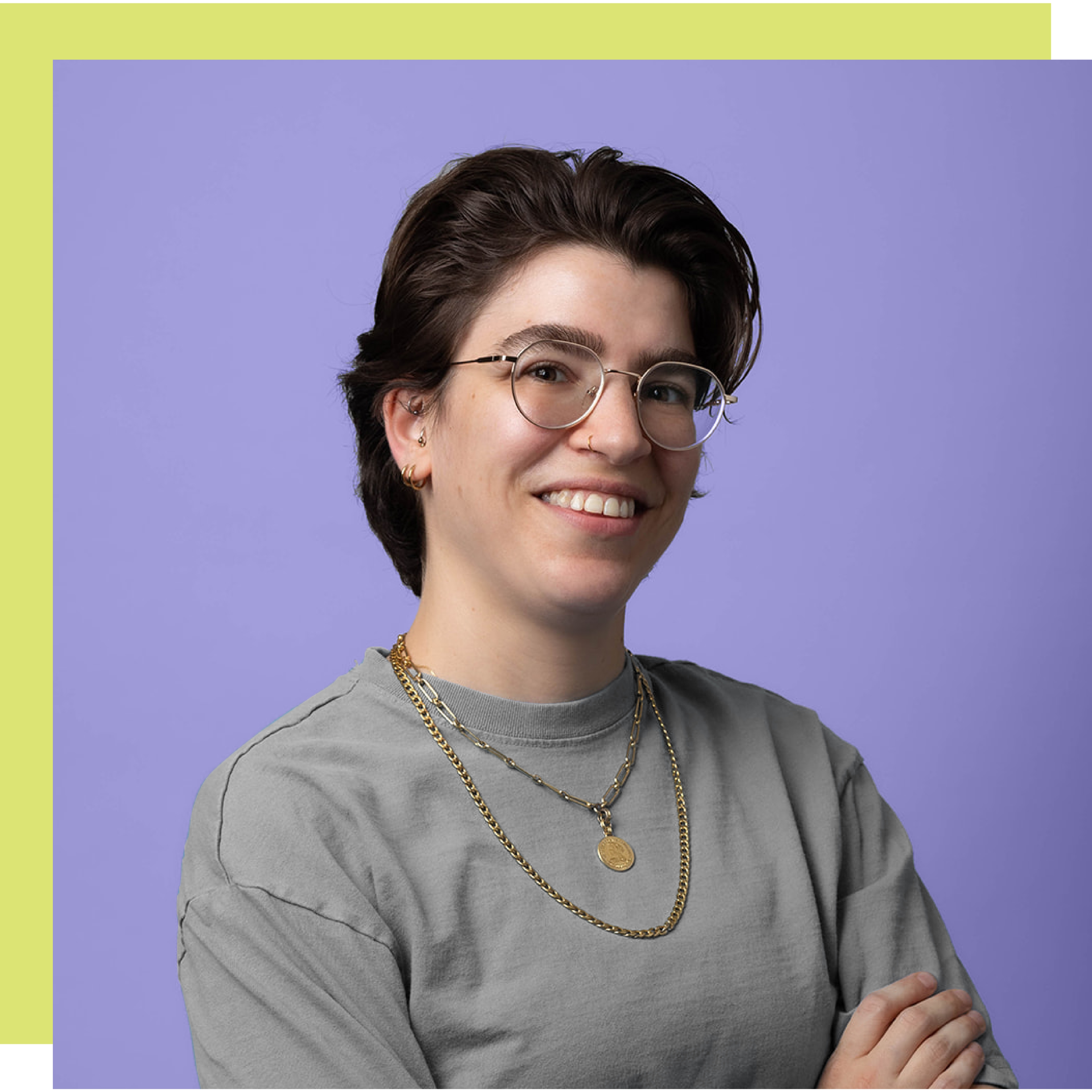 Smiling person with short dark hair, glasses, and a grey shirt against lavender background.